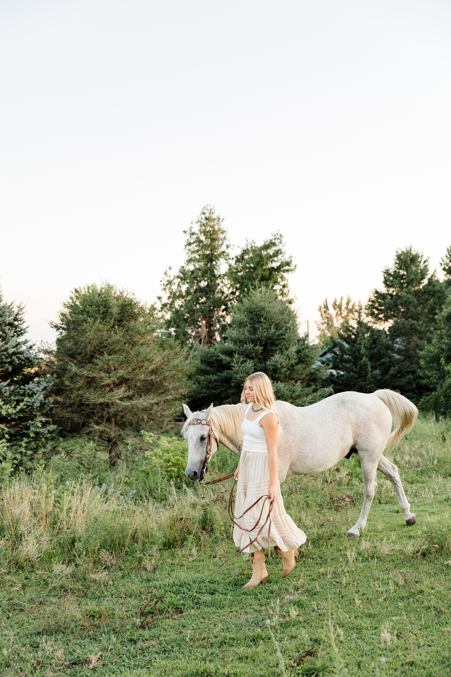 Emma, wearing a cream tank top and tan striped skirt, walks her tan colored horse in a pasture lined with pine trees during golden hour | CB Studio