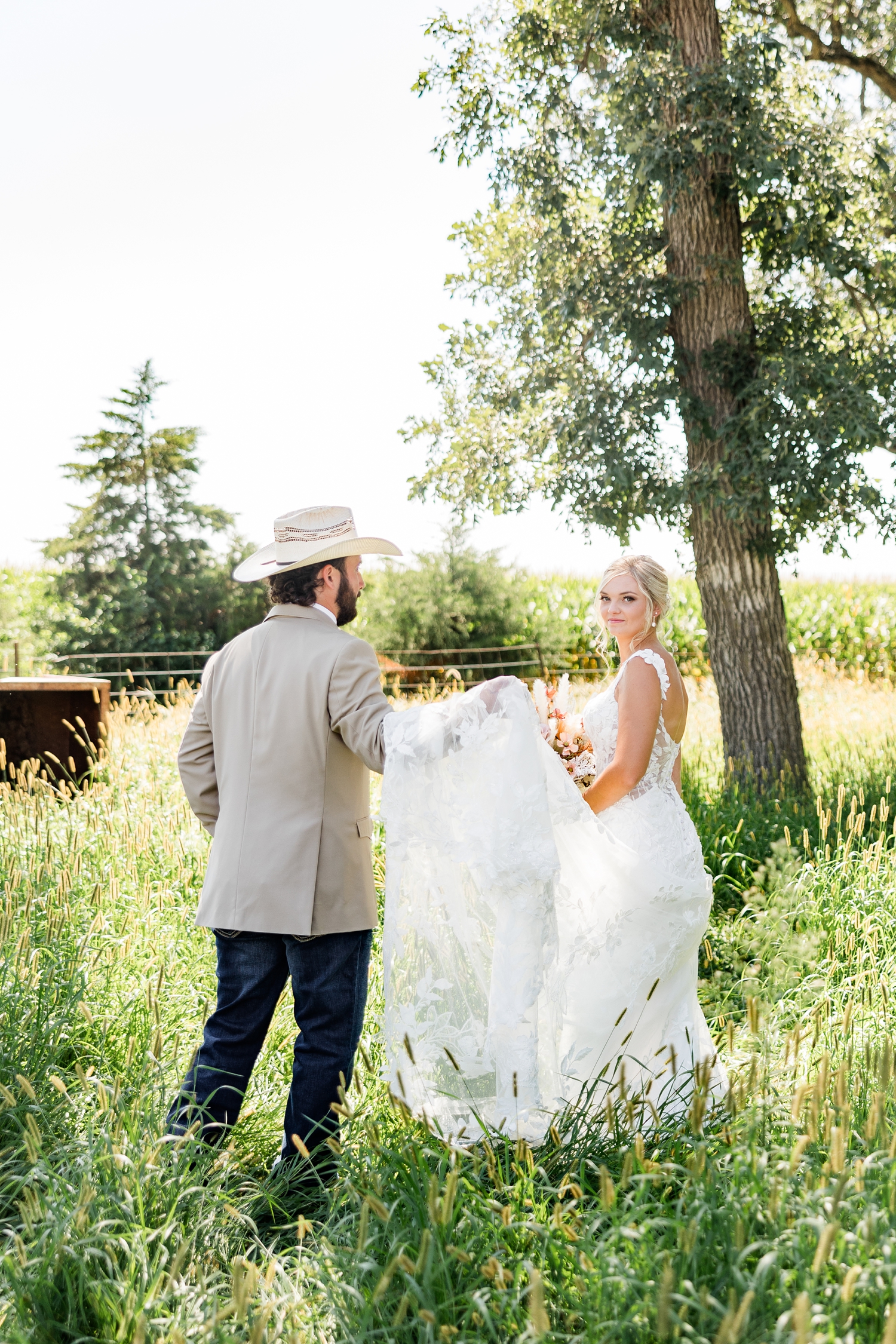 Jade walks out into a grass pasture and looks back as Dawson is behind her carrying her wedding gown train | CB Studio