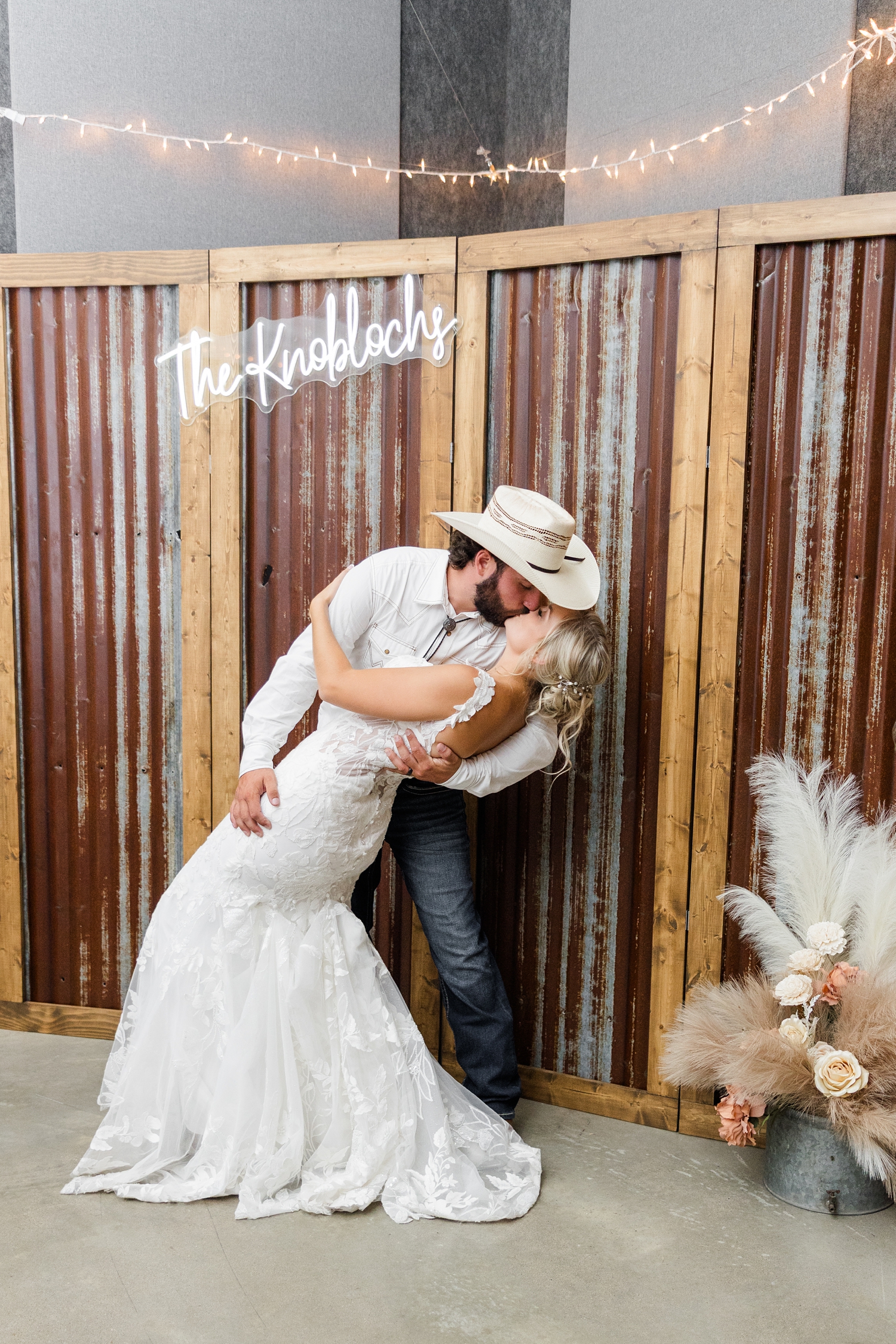 Dawson dips his bride for a kiss in front of their custom photo backdrop made of rustic steal panels and wood with a pampas grass and rose arrangement on the floor and a custom neon sign that reads in cursive "The Knoblochs" | CB Studio