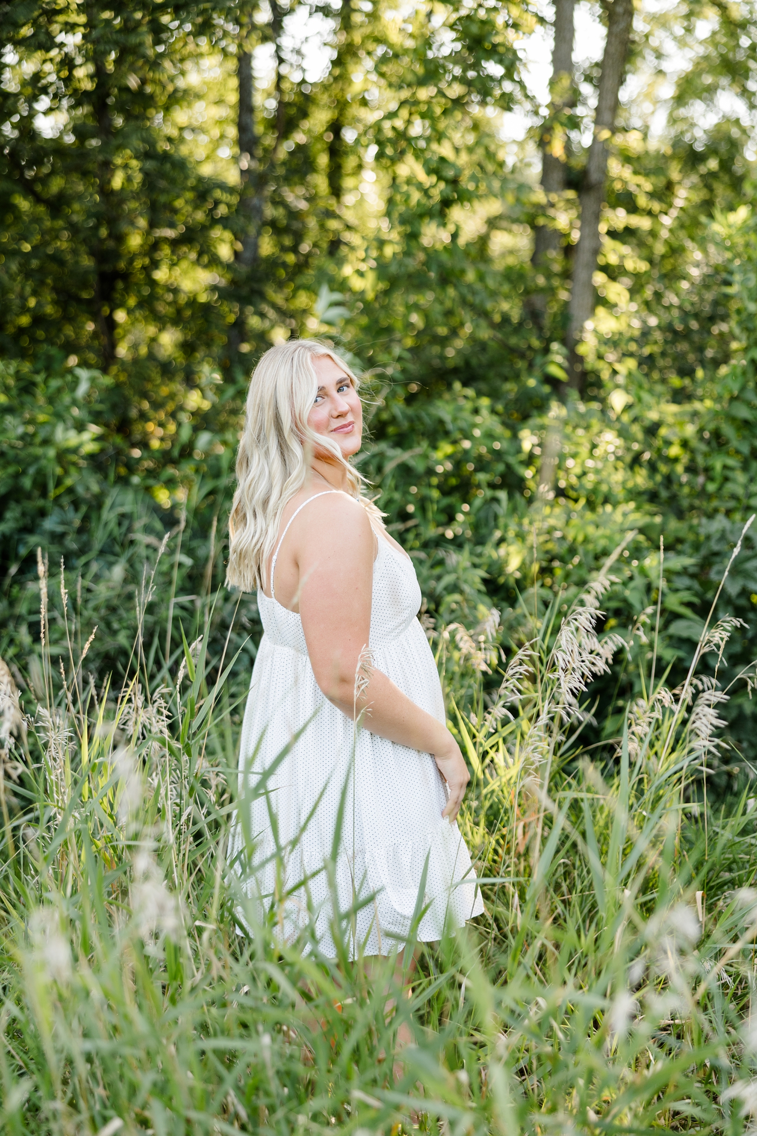 Ocean, wearing a white sundress, stands in a grassy meadow lined with trees in the background with her shoulder turned towards the camera as the breeze gently blows through her hair | CB Studio