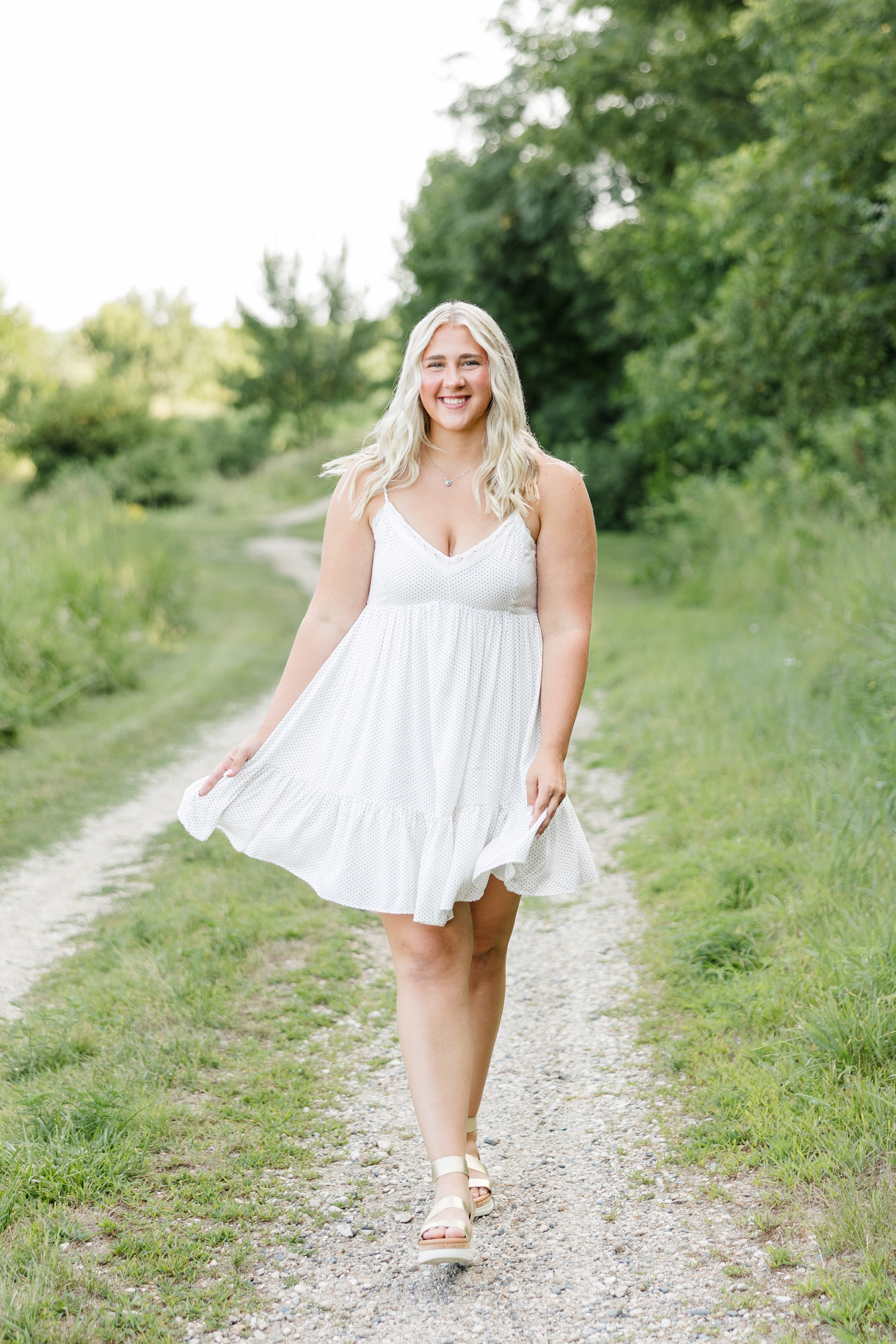 Ocean, wearing a white sundress, walks along a gravel path lined with trees at Water's Edge Nature Center | CB Studio