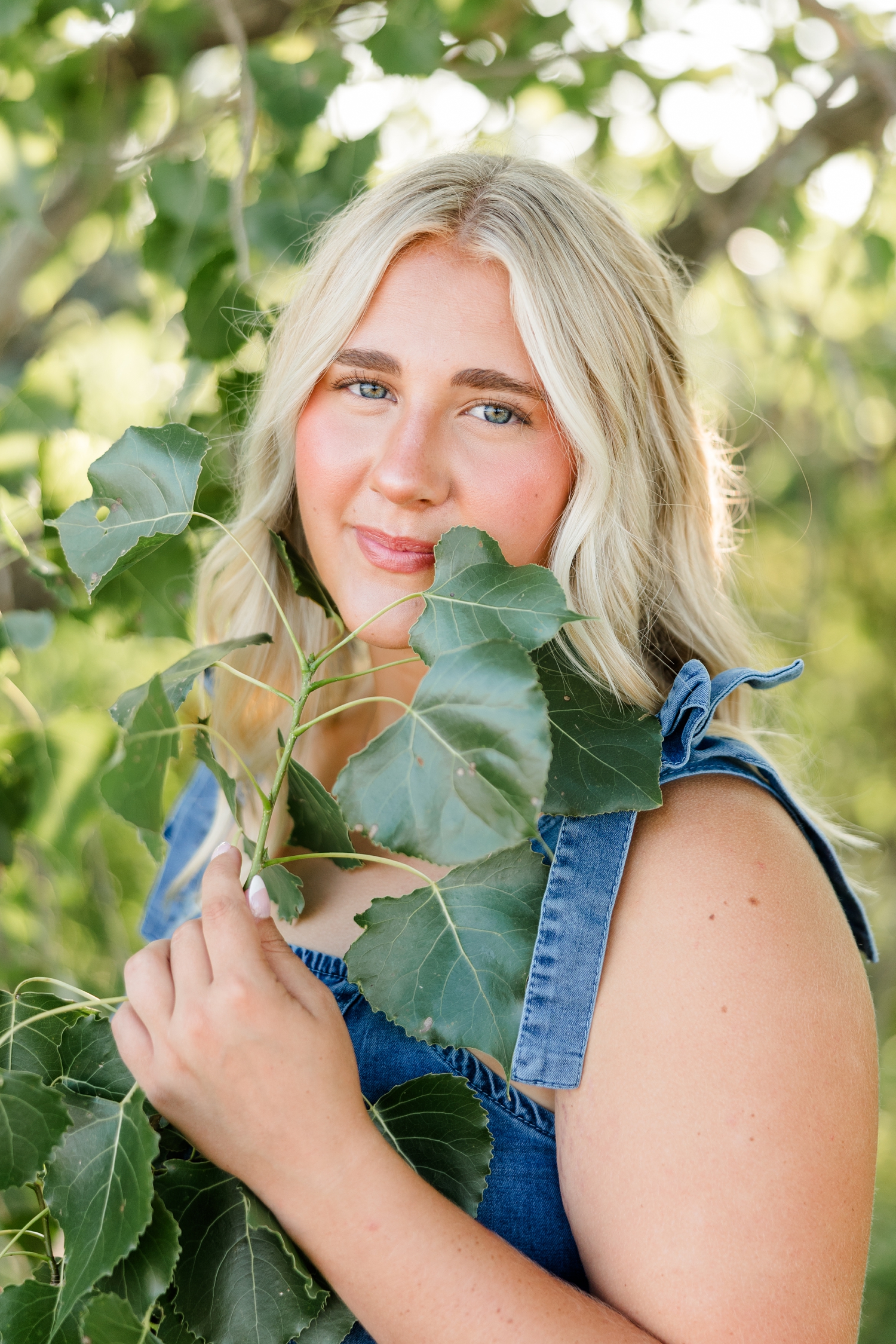 Ocean, wearing a denim dress, stands among the branches of a birch tree as she holds a small branch with leaves up to her chin | CB Studio