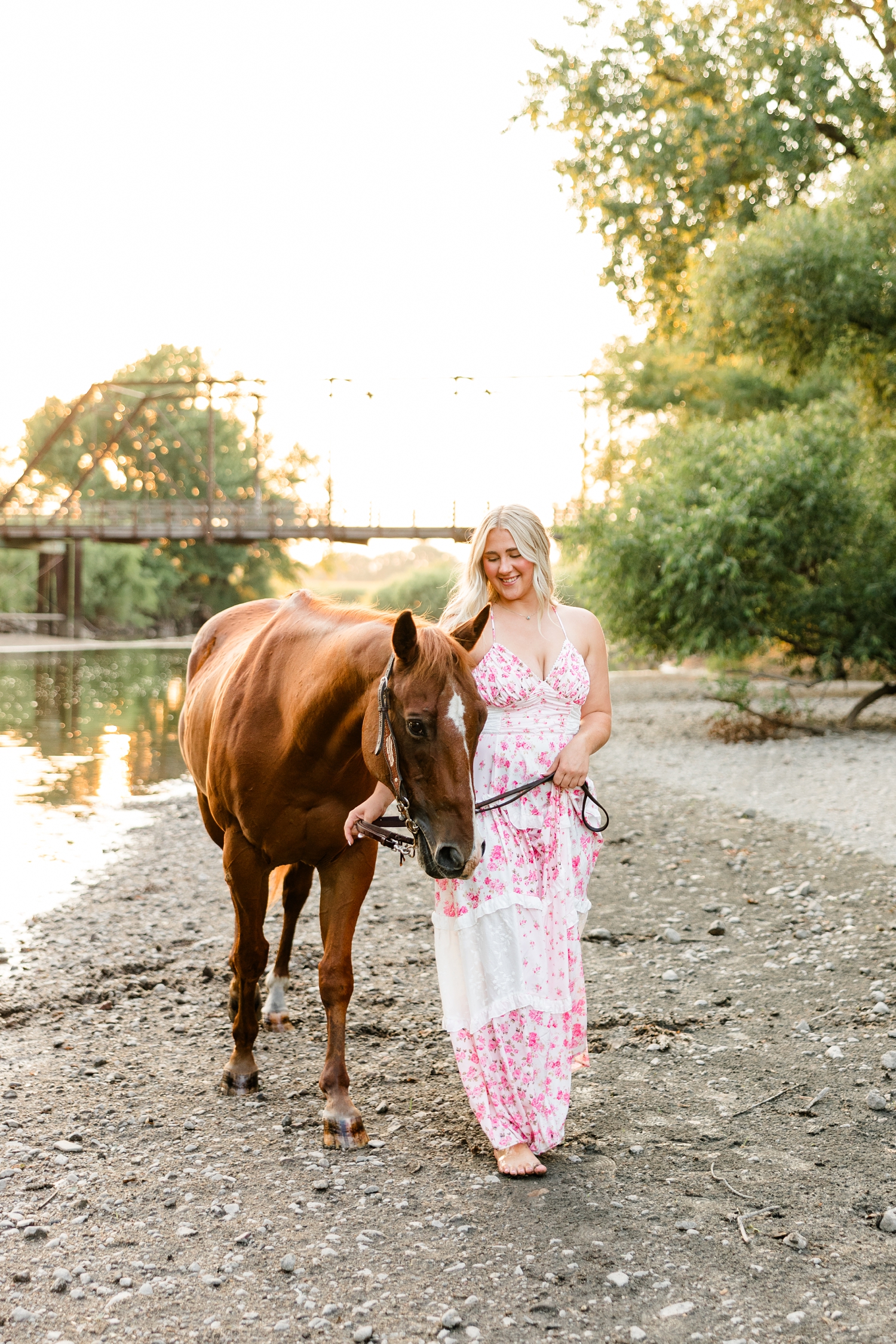 Ocean, wearing a white and pink floral maxi dress, walks her horse, Sparky, on a shallow river bank with an old trestle bridge in the background during golden hour | CB Studio