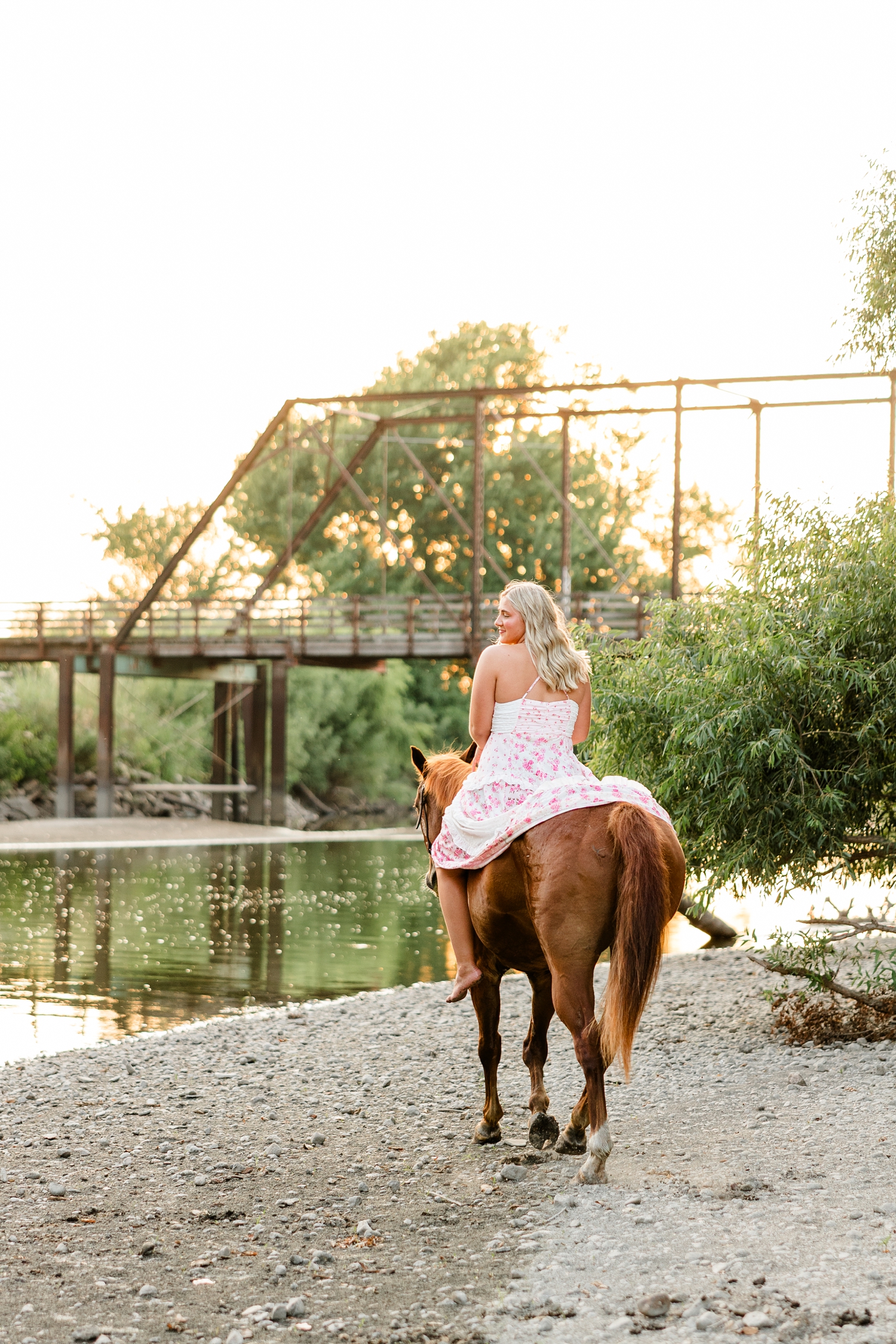 Ocean, wearing a white and pink floral maxi dress, rides on her horse, Sparky, off into the sunset on a shallow river bank with an old trestle bridge in the background | CB Studio
