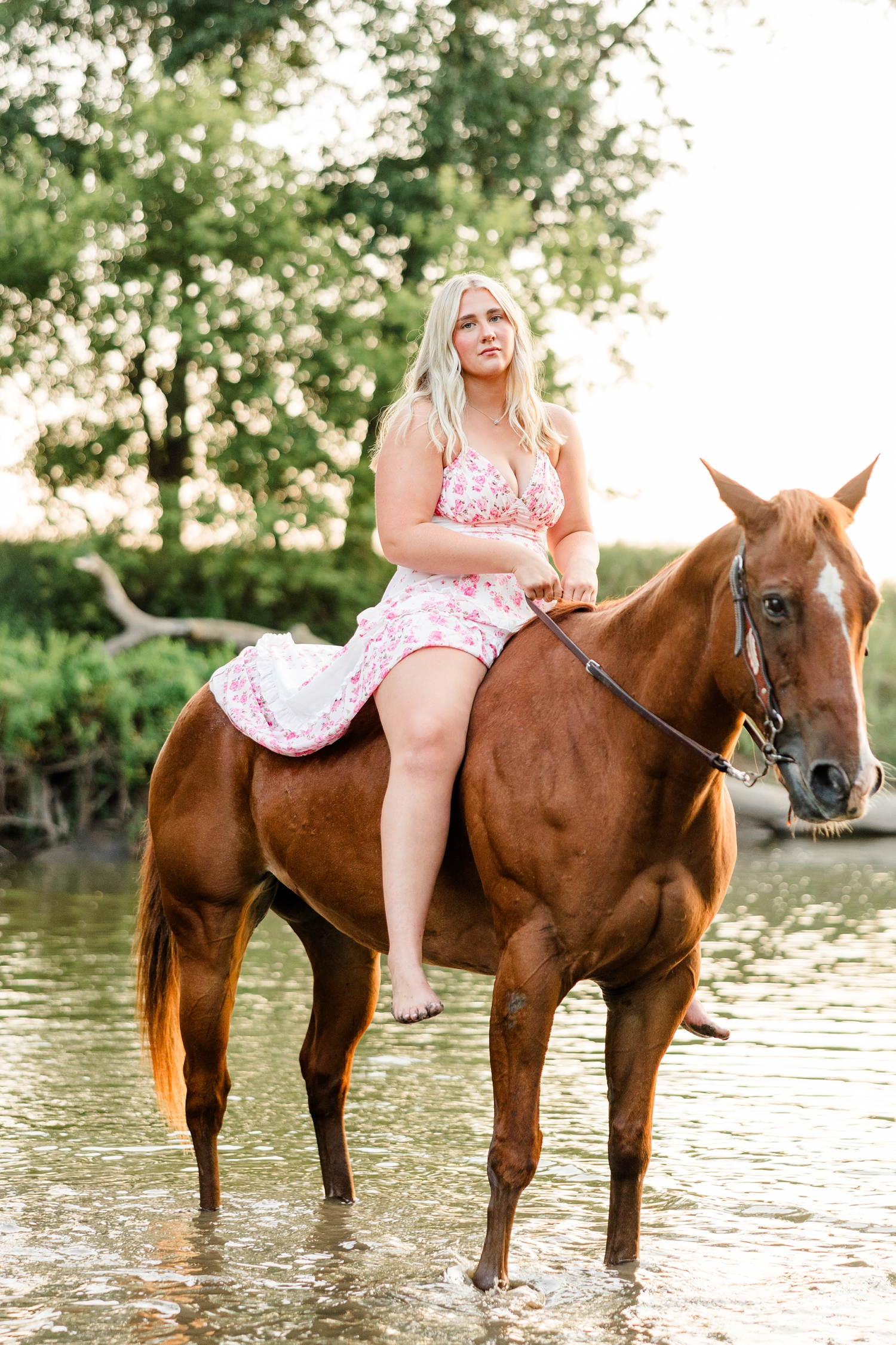 Ocean, wearing a white and pink floral maxi dress, sits on her horse, Sparky, in the middle of a shallow river bank during golden hour | CB Studio