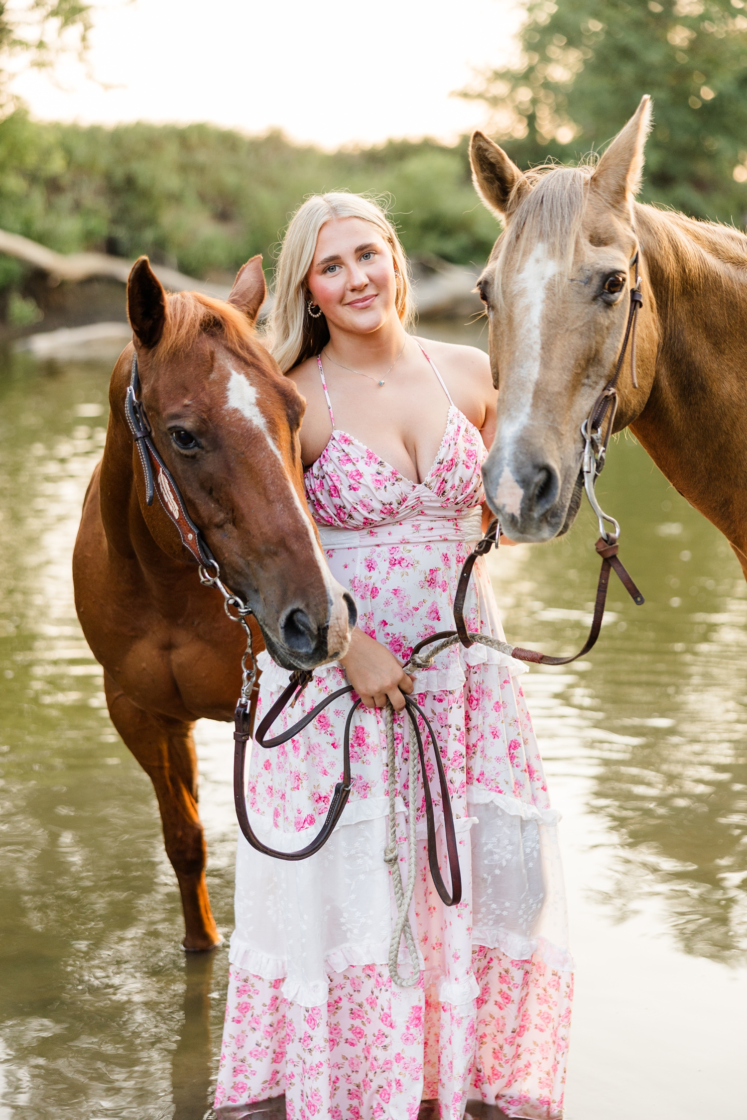 Ocean, wearing a white and pink floral maxi dress, stands in the middle of a shallow river with her two horses framing around her face | CB Studio