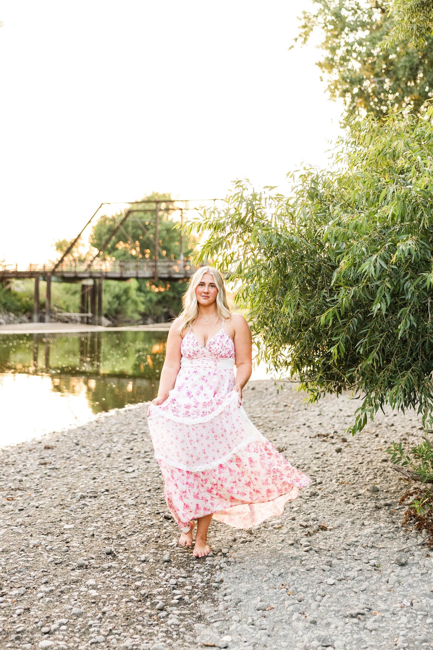 Ocean, wearing a white and pink floral maxi dress, walks along a shallow river bank at sunset with an old trestle bridge in the background | CB Studio