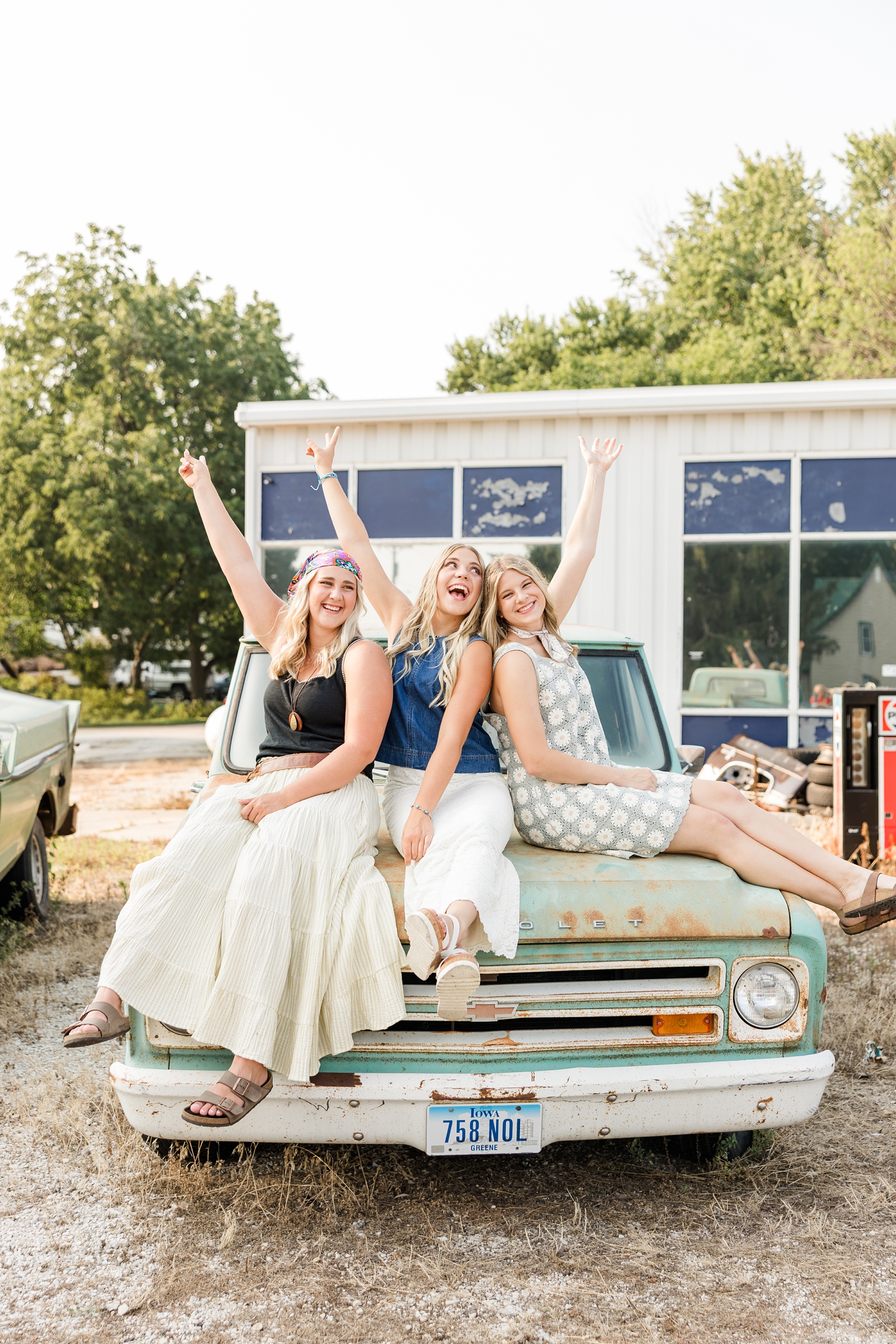 TEAM 27, dressed in boho, grunge looks, sit on the hood of an old rustic truck at a broken down gas station as they cheer with their hands in the air | CB Studio