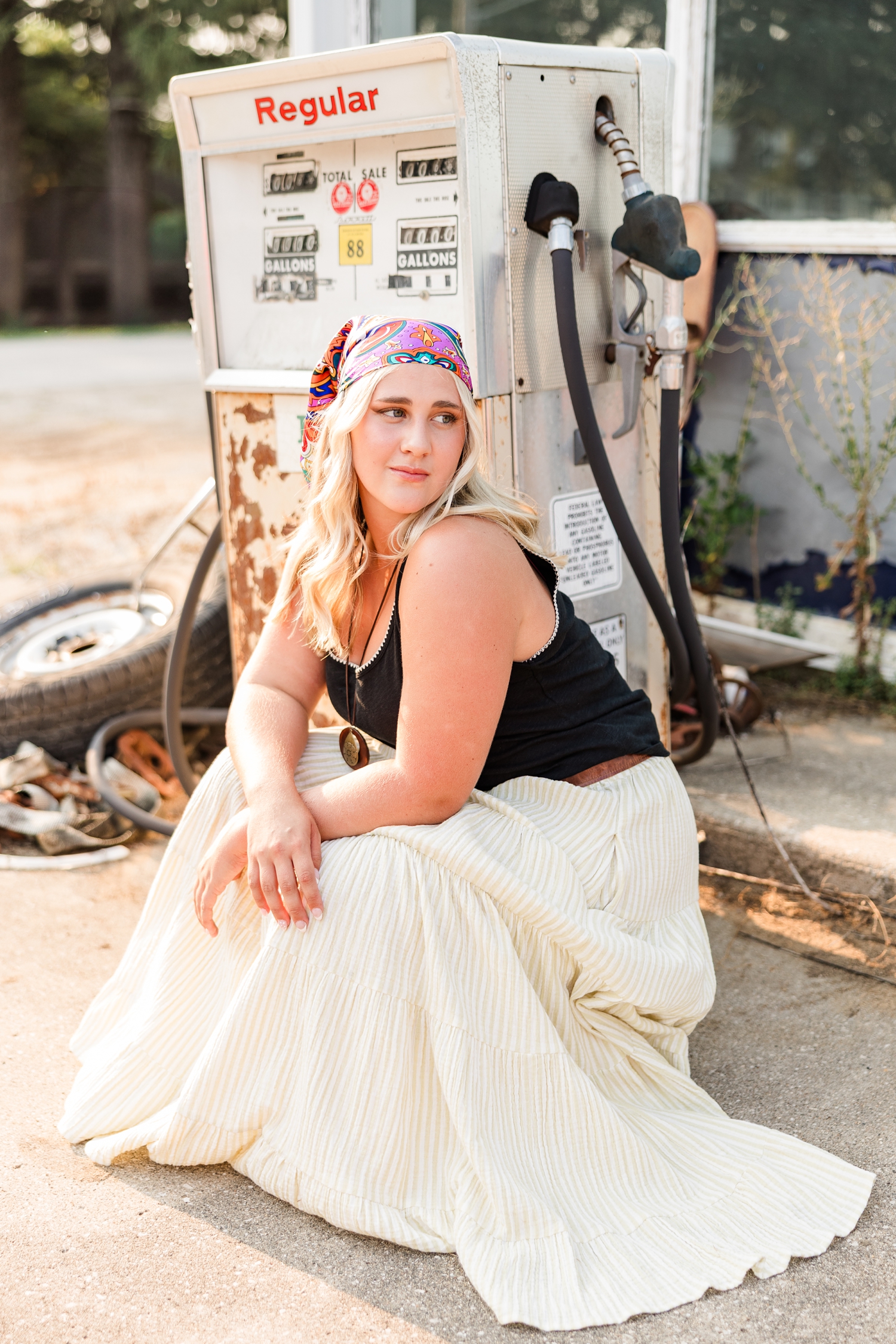 Ocean, dressed in a boho skirt and head scarf, squats in front of a broken down gas pump at an old gas station as she looks over her shoulder to the right | CB Studio