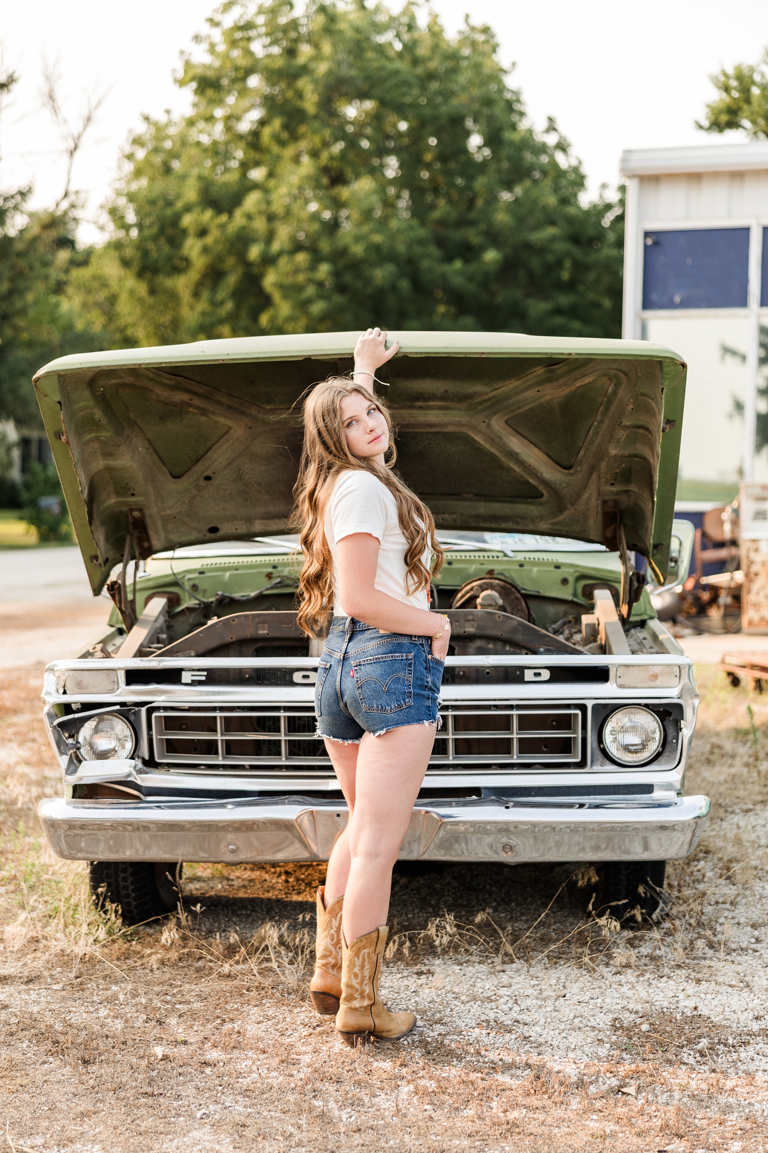 Mya, dressed in a grunge look, stand in front of an old Ford truck in front of an old broken down gas station as she holds the hood of the truck up and looks back over her shoulder | CB Studio