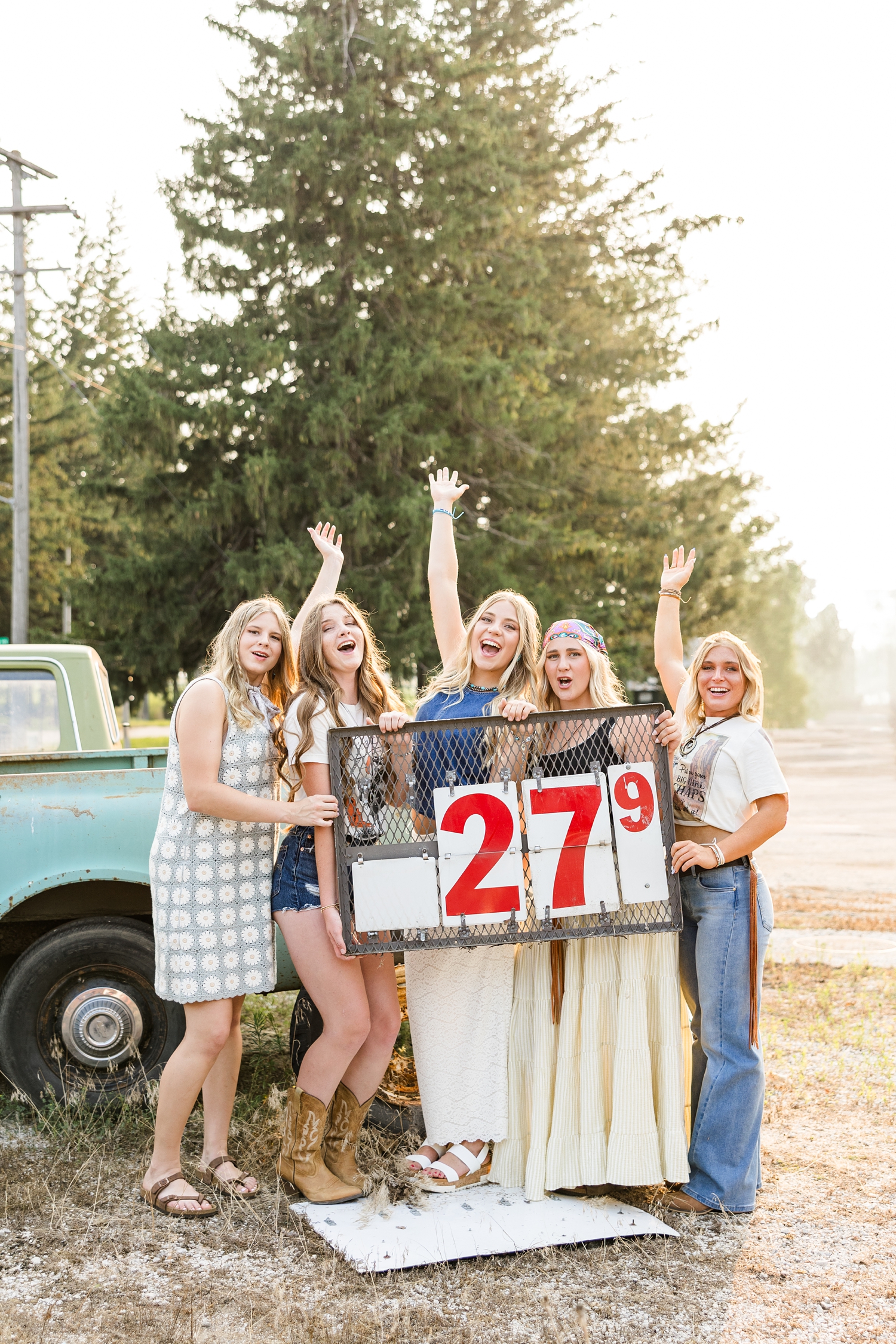 TEAM 27, dressed in boho, grunge looks, cheer as they hold an old gas price sign with 27 cents listed at an old car lot | CB Studio