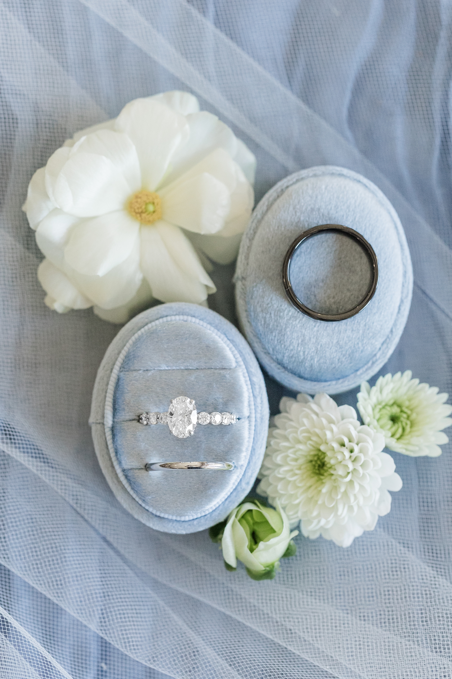 Cassie's oval engagement ring with round diamond band and platinum wedding band sit in a light blue velvet box with Ryan's black wedding band sits atop the velvet ring box top all surrounded by florals sitting atop a light blue fabric and veil | CB Studio