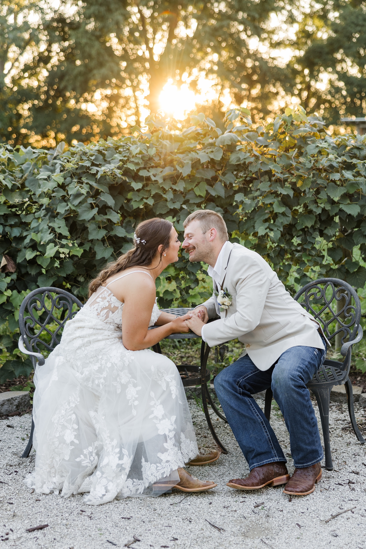 Ryan and Cassie sit at a cast iron cafe table opposite each other, holding hands, and slowing going in for a kiss amongst the vineyards at River Valley Orchards at sunset | CB Studio