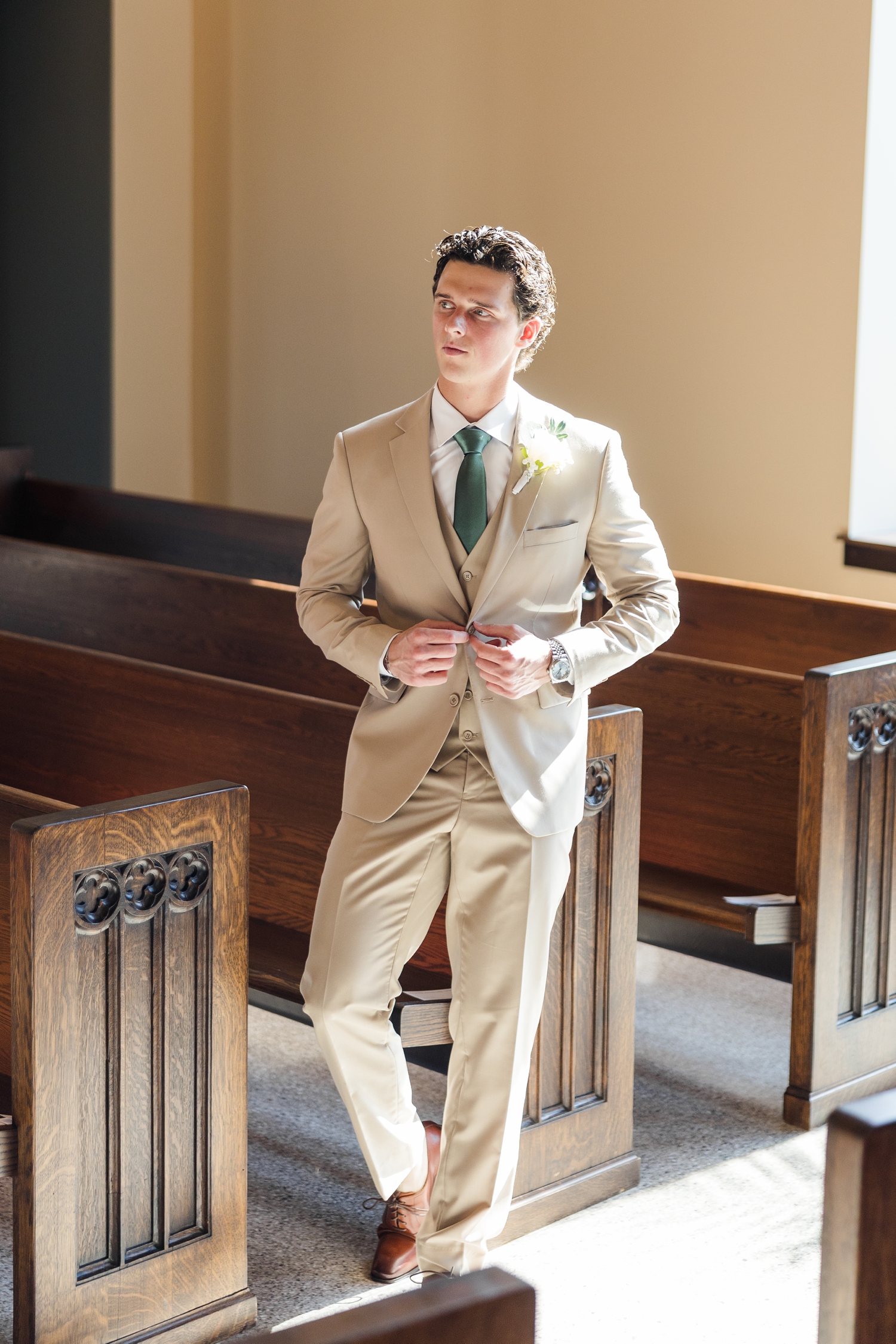 Groom buttons his jacket in the sunlight of a large window of the Holy Trinity Catholic Church in Fort Dodge, IA | CB Studio