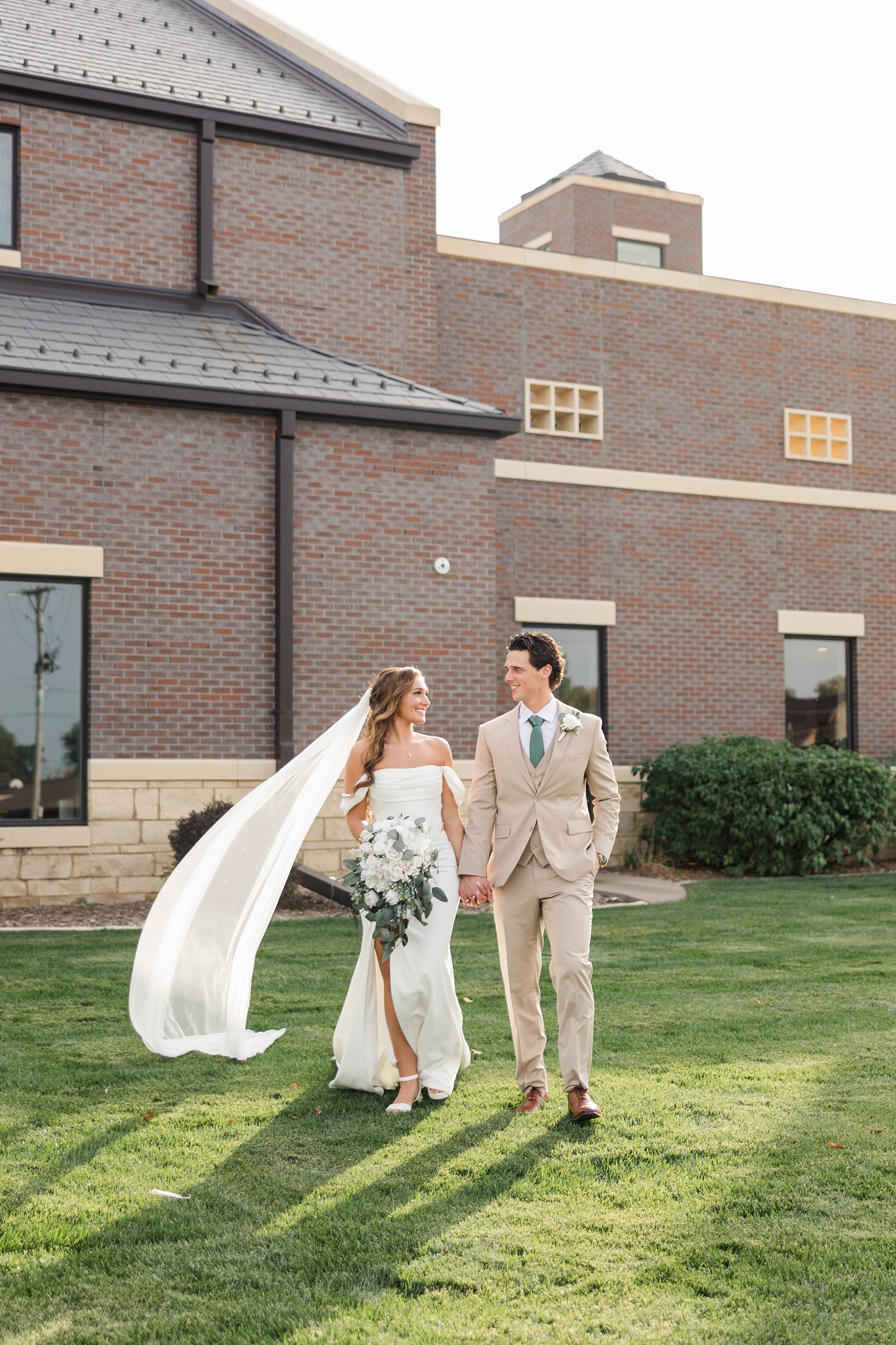 Bride and groom walk across the lawn in front of the Holy Trinity Catholic Church in Fort Dodge, IA | CB Studio