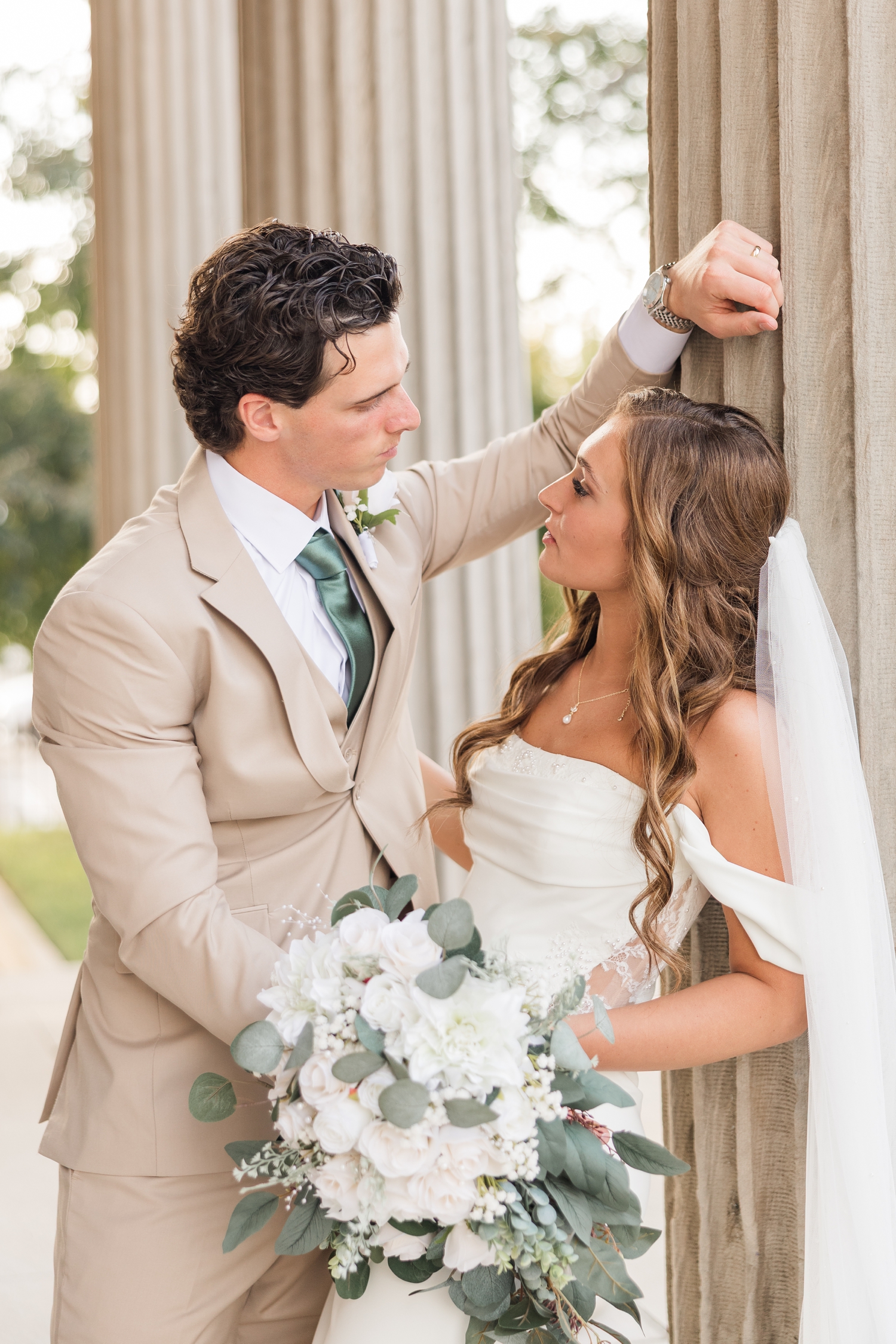 Bride and groom lean against the column of a historic building in Fort Dodge, IA as they both look at each other | CB Studio
