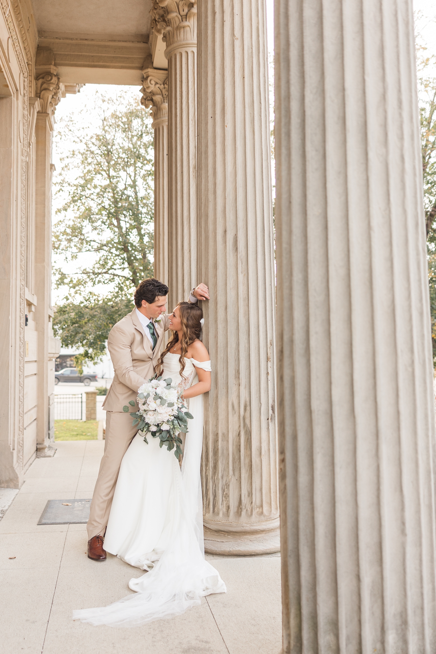 Bride and groom lean against the column of a historic building in Fort Dodge, IA as they both look at each other | CB Studio