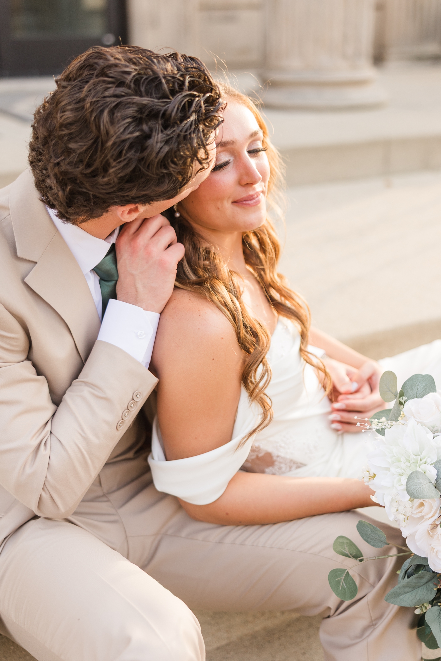 Bride and groom sit together on the steps of a historic building in Fort Dodge, IA as the groom kisses the bride's cheek at sunset | CB Studio