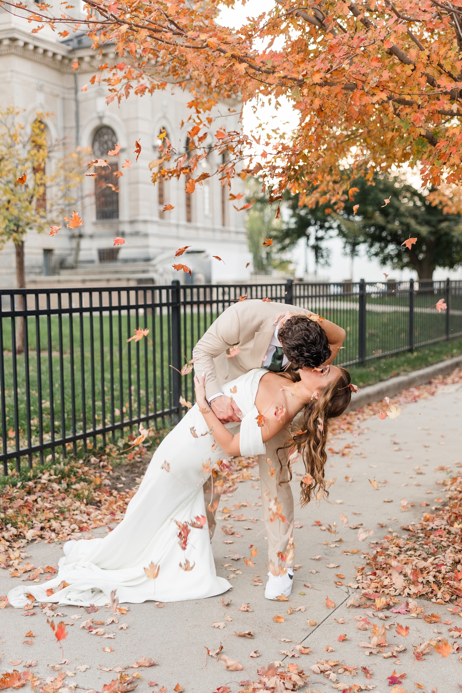 Tyler dips his new bride for a kiss on a sidewalk in Fort Dodge, IA under a tree at peek fall as leaves are falling down around them | CB Studio