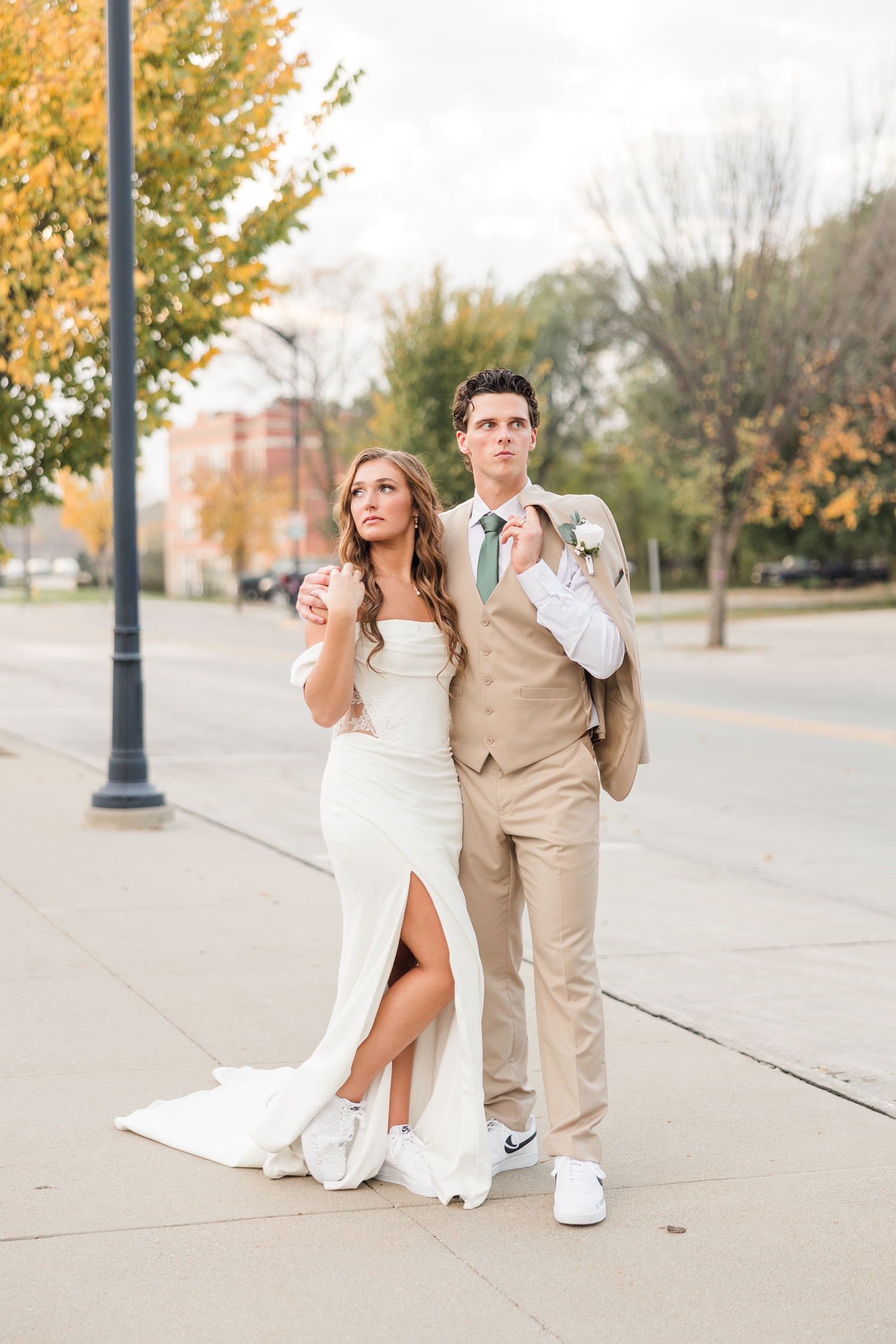 Bride and groom stand along a fall tree lined street in Fort Dodge, IA as they model pose and look off into opposite directions | CB Studio