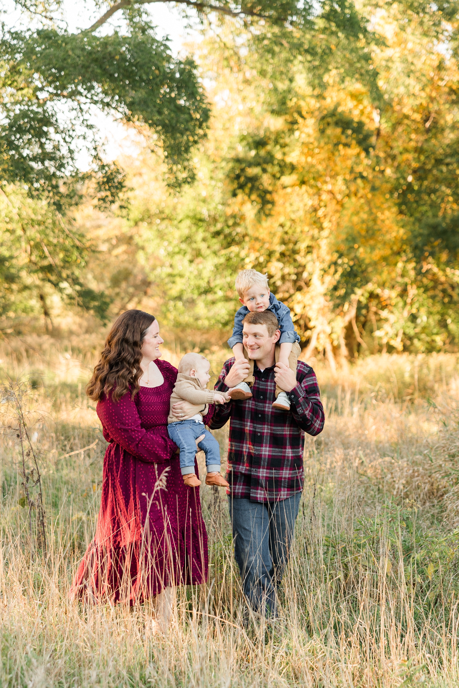 The Fischels family walks through a fall grassy pasture lined with oak trees as sun shines through the leaves while they smile at each other | CB Studio