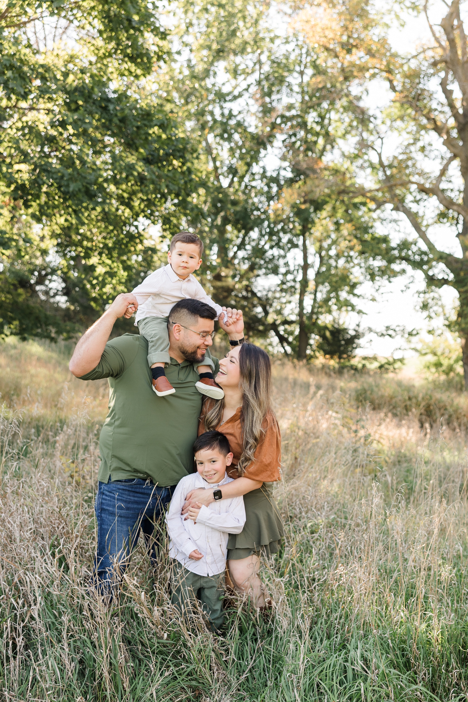 The Hernandez family stand and embrace in a fall grassy pasture lined with oak trees as the littlest boy sits on dad's shoulders and mom and dad smile at each other | CB Studio