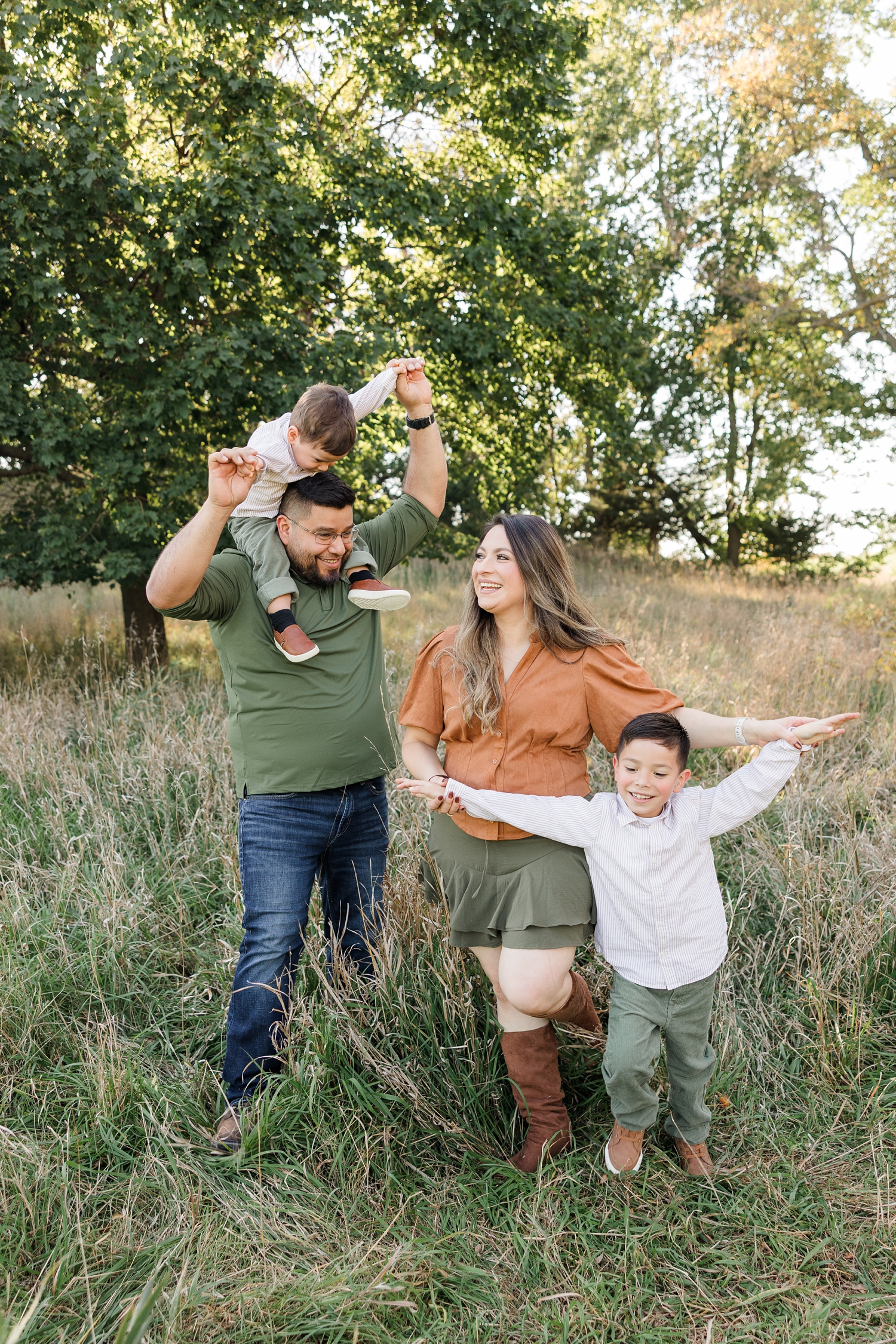 The Hernandez family walks through a fall grassy pasture lined with oak trees as they play "airplanes" with their two little boys | CB Studio